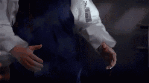a man in a lab coat and blue apron is standing in a dark room with his hands outstretched .