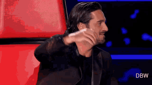 a man in a black jacket is sitting in a red chair and covering his face with his hand .