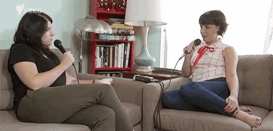 two women are sitting on a couch talking into microphones