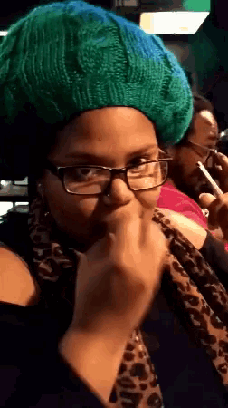 a woman wearing glasses and a green hat holds her hand to her face