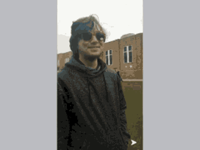 a young man wearing sunglasses and a hoodie stands in front of a brick building