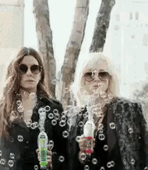 two women wearing sunglasses are blowing soap bubbles in front of trees .