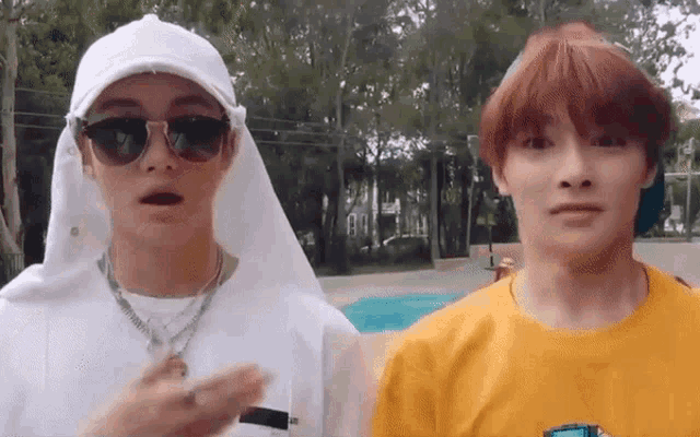 two young men are standing next to each other in a park . one of the men is wearing sunglasses and a white hat .