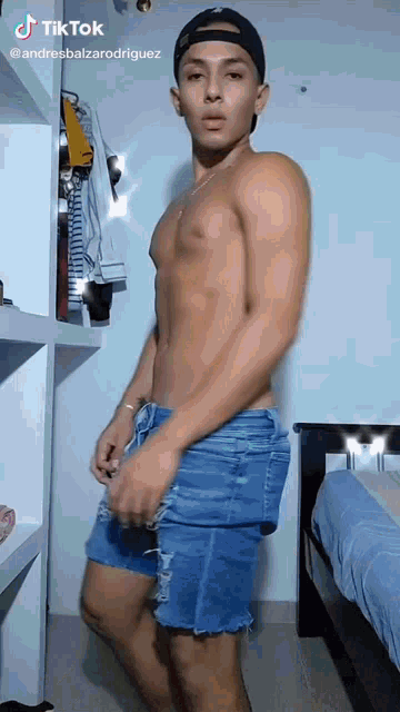 a shirtless man wearing denim shorts and a hat is standing in a bedroom .