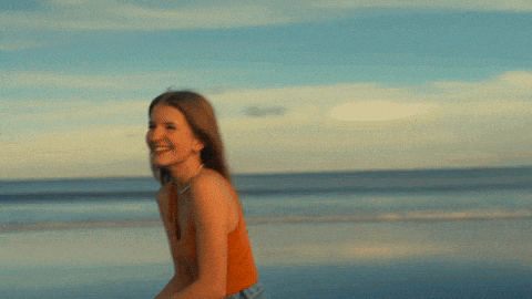a woman in an orange tank top and jeans is running on the beach
