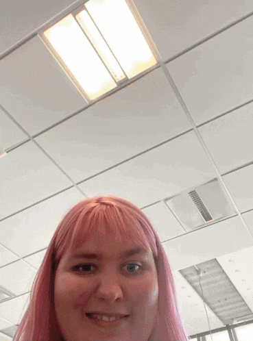 a woman with pink hair smiles in front of a white ceiling