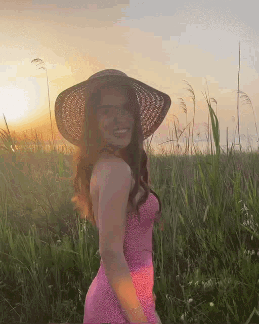 a woman in a pink dress and straw hat stands in a field at sunset