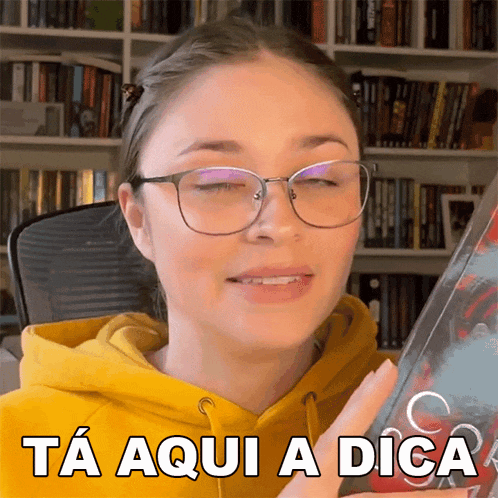 a woman wearing glasses and a yellow hoodie is holding a book and says ta aqui a dica