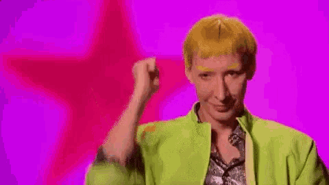 a woman in a green jacket is standing in front of a pink background and making a funny face .