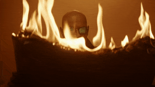 a bald man wearing glasses looks out from behind a pile of flames