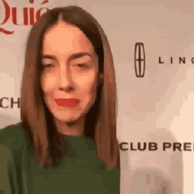 a woman wearing a green sweater and red lipstick is standing in front of a wall with a lincoln logo .