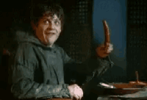a man is sitting at a table holding a sausage and making a face .