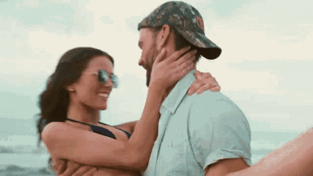a man is holding a woman in his arms on the beach and the woman is wearing sunglasses .