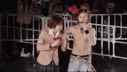 a man without a shirt is singing into a microphone while a girl in a school uniform holds his arm .