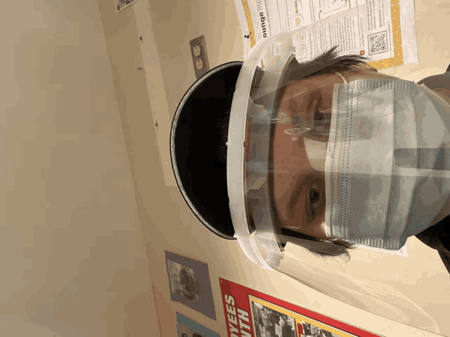 a person wearing a face shield and a mask stands in front of a poster that says " dyees with "