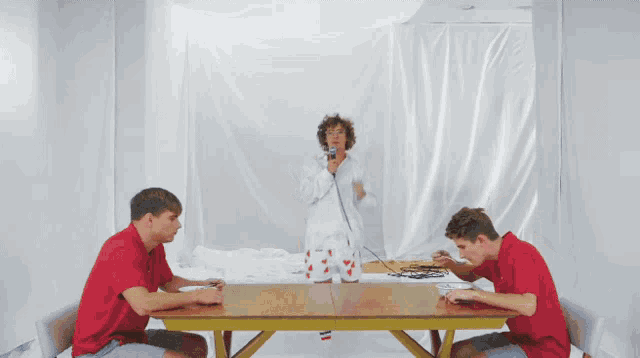 two men in red shirts sit at a table with a woman in a white shirt standing in the background