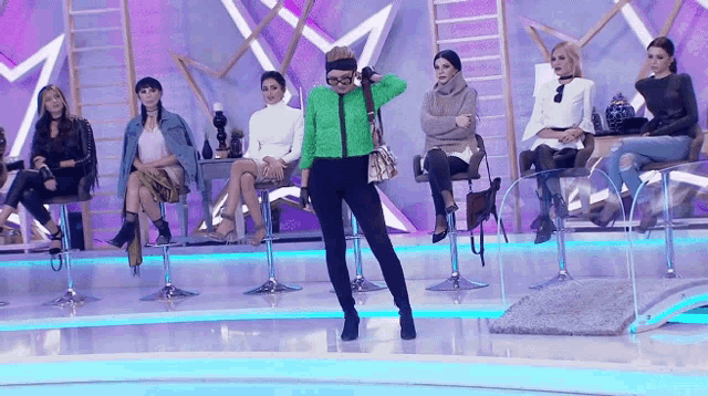 a woman in a green sweater stands in front of a group of women sitting on stools