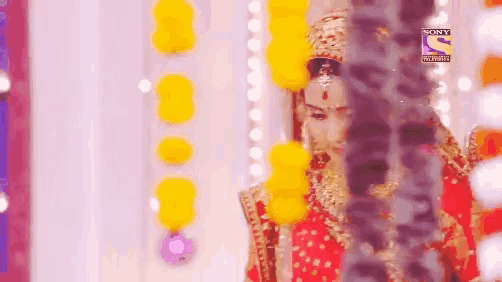 a bride in a red and gold wedding dress is standing in front of a wall with yellow flowers hanging from it .