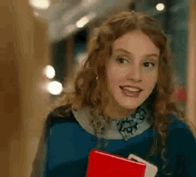 a woman with curly hair is holding a red book and smiling while walking down a hallway .