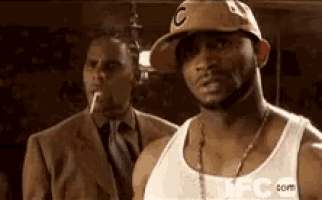 a man in a hat is smoking a cigarette next to another man in a suit