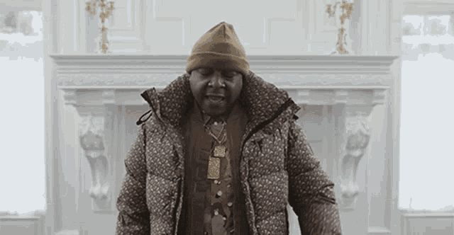 a man wearing a beanie and a jacket is standing in front of a fireplace .