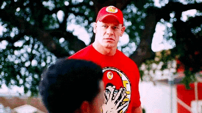 a man wearing a red shirt and a red hat is standing next to another man covering his face .