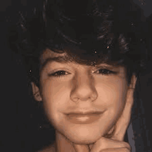 a young boy with curly hair is smiling and holding his hand to his chin .