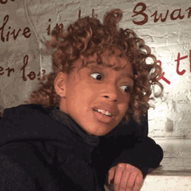 a girl with curly hair is standing in front of a brick wall that says live in