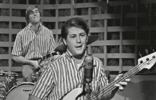 a man is singing into a microphone while playing a bass guitar in front of a drum set .