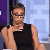a woman wearing glasses is sitting at a table with her hand on her chin and making a funny face .