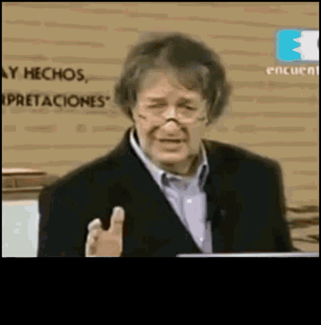a man in a suit and glasses is giving a speech in front of a sign that says " ay hechos "