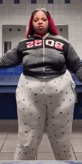 a woman with pink hair is standing in a bathroom wearing pajamas and a hoodie with the number 2008 on it .