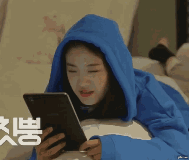 a woman in a blue hoodie is laying on a couch looking at a tablet .