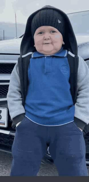 a little boy wearing a hoodie and a beanie stands in front of a car with a license plate that says 3