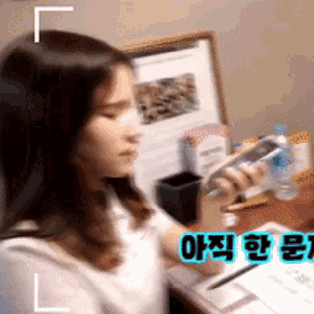 a woman is sitting at a desk holding a cell phone in front of a board with chinese writing on it