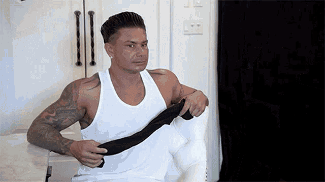a man wearing a white tank top is sitting in a chair holding a black tie .