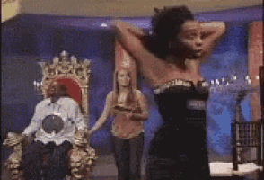 a woman in a black dress is standing in front of a man in a gold chair
