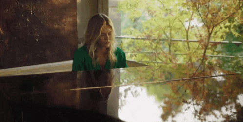 a woman in a green shirt sits at a piano in front of a window