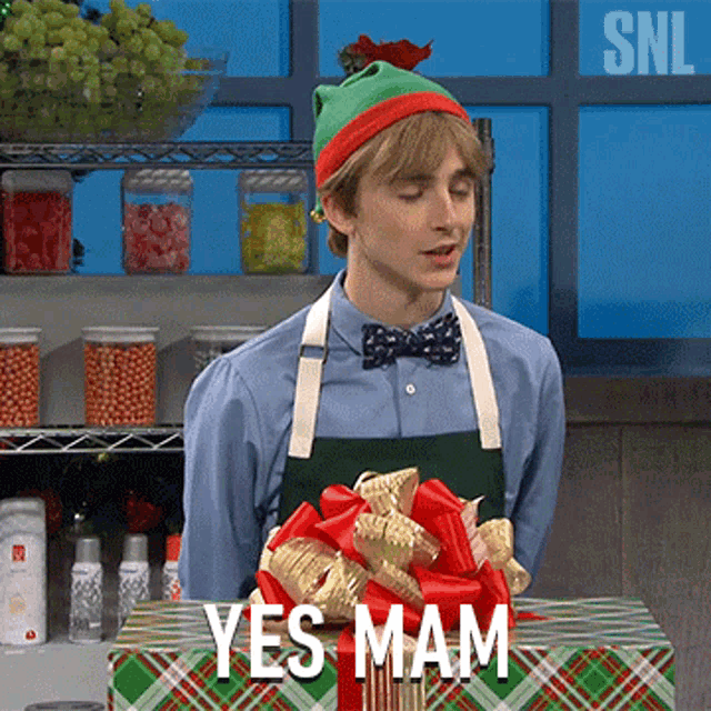 a man wearing an elf hat and apron is standing in front of a gift box that says " yes mam "