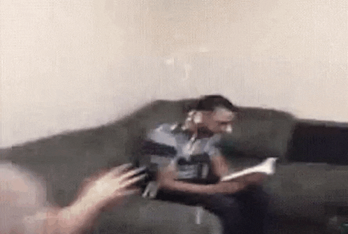 a man is sitting on a couch reading a book and talking on a phone .