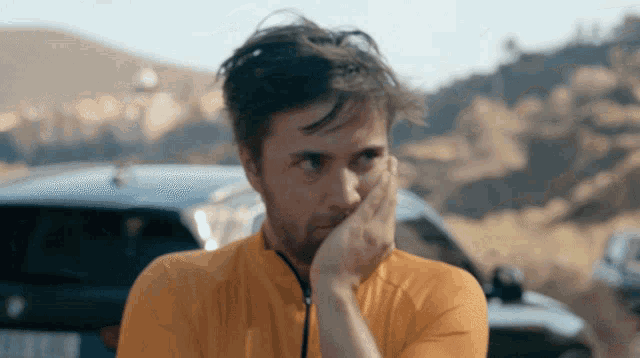 a man covering his mouth with his hand in front of a car with a license plate that starts with the letter c
