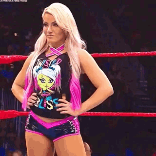 a woman in a pink and black shirt is standing in a wrestling ring with her hands on her hips .