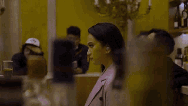 a woman in a pink jacket is sitting at a table in front of a yellow wall