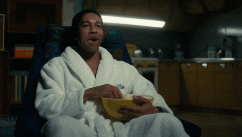 a man in a bathrobe is sitting in a chair and eating popcorn