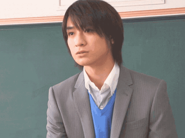 a man wearing a suit and a blue vest stands in front of a blackboard