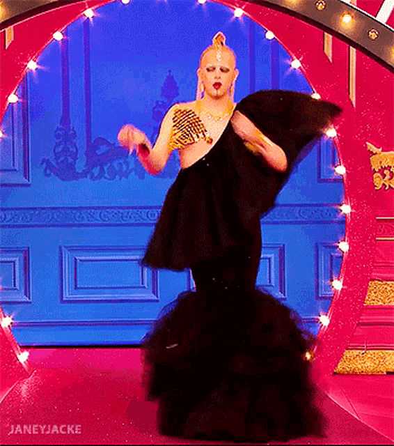 a drag queen in a black dress is dancing in front of a pink archway .