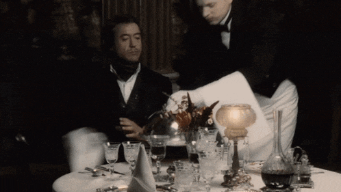 a man in a tuxedo sits at a table while a waiter pours a glass of wine