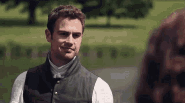 a man in a vest stands in a park looking at another man