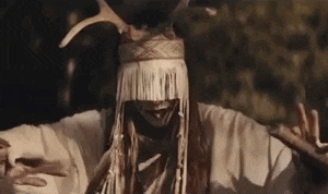 a man in a native american costume with antlers on his head is standing in the dark with his arms outstretched .