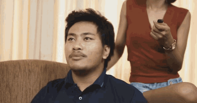 a man with a mustache sits on a couch while a woman sits behind him holding a lighter